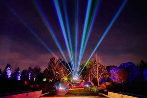 Colorful laser beams illuminate a dark sky over a park, with trees and glowing spheres in the foreground.