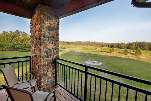 A balcony view featuring stone pillars, two chairs, and a scenic landscape with green fields and trees.