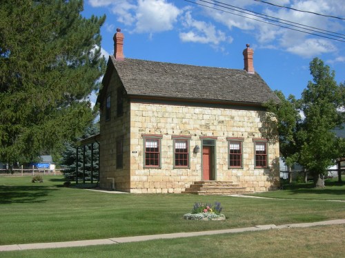 A historic stone house with a sloped roof, red door, and surrounded by green grass and trees under a blue sky.