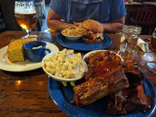 A table with a plate of ribs, macaroni, cornbread, a burger, and a beer in a casual dining setting.