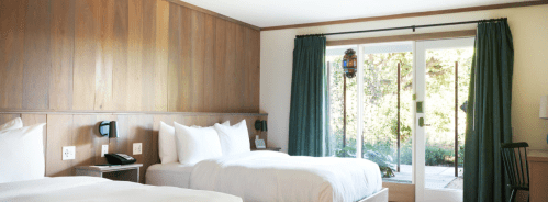 A cozy hotel room with two beds, wooden walls, and a sliding door leading to a patio with greenery outside.
