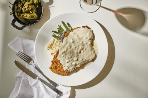 A plate of fried chicken topped with creamy gravy, served with green beans and a side of mashed potatoes.