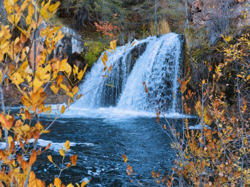 A serene waterfall cascades into a blue pool, surrounded by autumn leaves in vibrant yellow and orange.