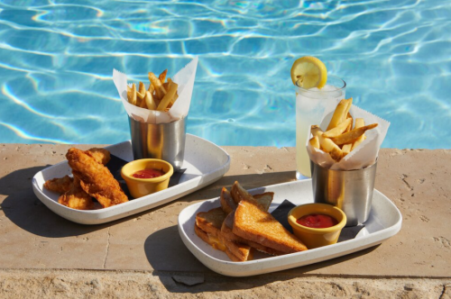 Two trays of food by a pool: one with fried chicken and fries, the other with grilled cheese and fries, plus a drink.