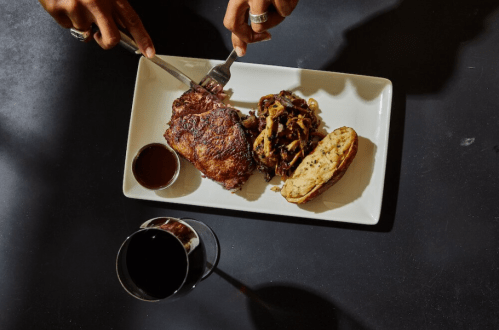 A plate with sliced meat, a bun with toppings, and a glass of dark beverage on a black table.
