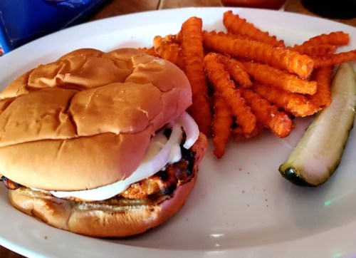 A plate with a burger topped with onions, served with sweet potato fries and a pickle on the side.