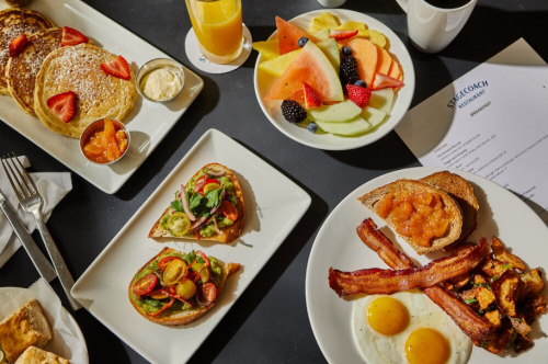 A vibrant breakfast spread featuring pancakes, fruit, toast with toppings, bacon, and eggs on a table.