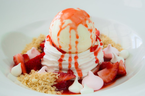 A dessert featuring a scoop of vanilla ice cream on whipped cream, topped with strawberry sauce and surrounded by strawberries and meringues.