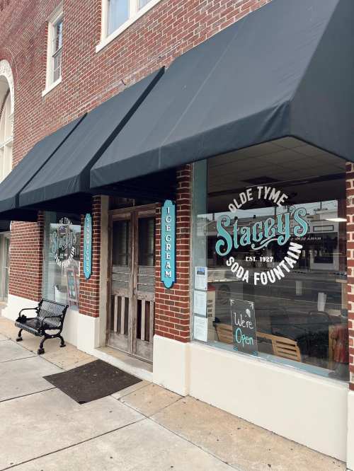 Exterior of Stacey's Olde Tyme Soda Fountain, featuring a brick facade, large windows, and a black awning. "We're Open" sign visible.
