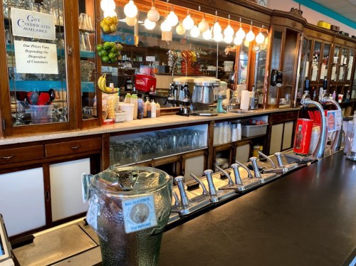 A vintage diner counter with a soda fountain, glassware, and a wooden backdrop featuring hanging lights and decor.