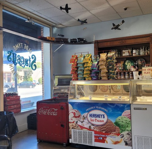 Interior of an ice cream shop with a vintage Coca-Cola cooler, colorful snacks, and model airplanes on display.