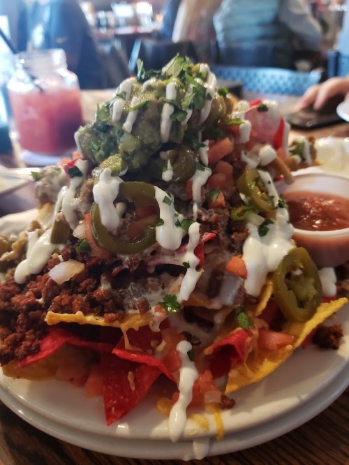A plate of loaded nachos topped with guacamole, jalapeños, diced tomatoes, sour cream, and a side of salsa.