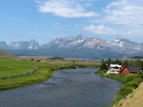 A serene river flows through green fields, with mountains in the background and a few houses along the bank.
