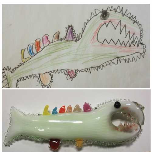 A child's drawing of a green fish with sharp teeth above a glass fish sculpture with colorful fins and a similar design.