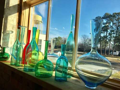 A collection of colorful glass vases in various shapes, displayed on a windowsill with sunlight streaming in.