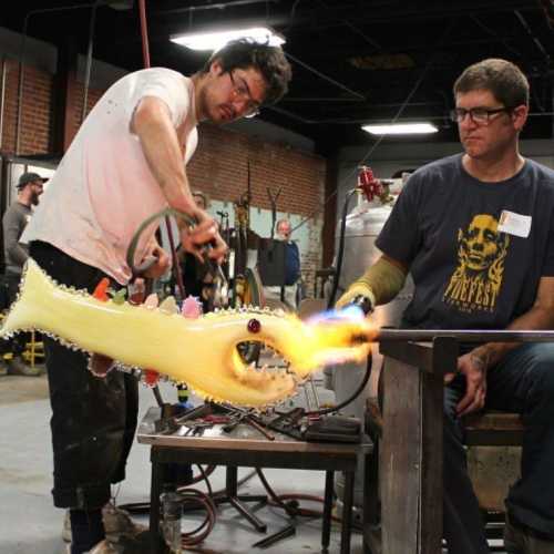 Two glassblowers work on a colorful glass sculpture in a workshop, with tools and equipment visible around them.