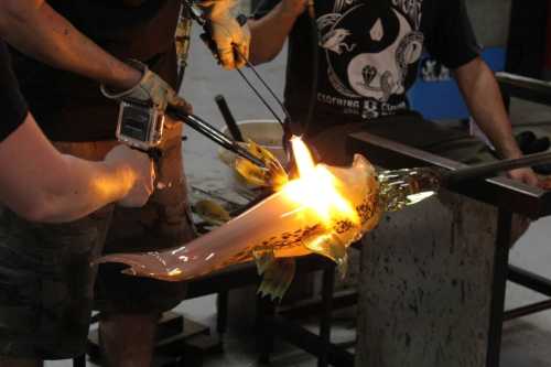 A glass artist shapes a glowing fish sculpture using tools, with a team assisting in the glassblowing process.