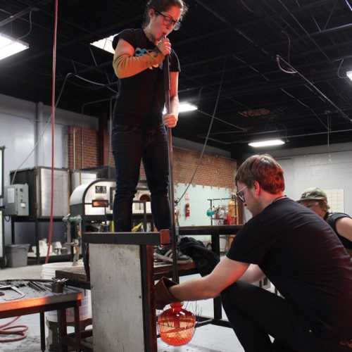 A glassblower shapes molten glass while a colleague assists in a workshop setting.