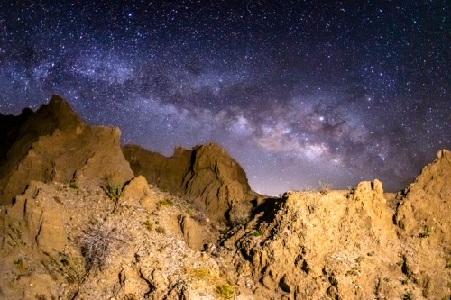 A starry night sky filled with the Milky Way over rugged desert terrain and rocky formations.