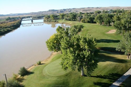 Aerial view of a lush green golf course beside a calm river, with trees and hills in the background.