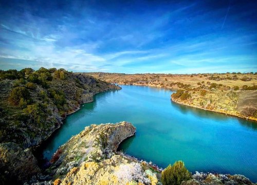 A serene river winding through rocky terrain under a clear blue sky, surrounded by lush greenery.