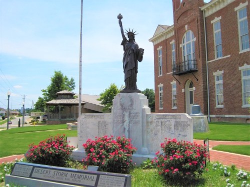Statue of Liberty replica on a memorial pedestal, surrounded by flowers, near a brick building and gazebo.