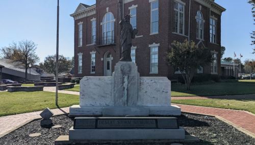 Statue of a soldier on a pedestal in front of a historic brick building, surrounded by grass and pathways.