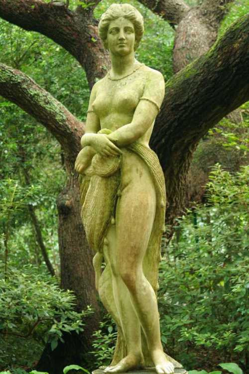 A weathered statue of a woman standing gracefully, draped in fabric, surrounded by lush greenery and trees.