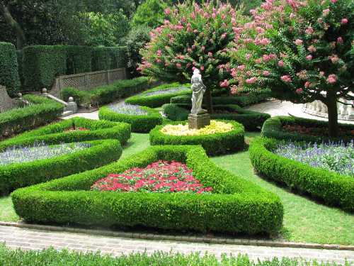 Lush garden with neatly trimmed hedges, colorful flower beds, and a statue surrounded by blooming trees.