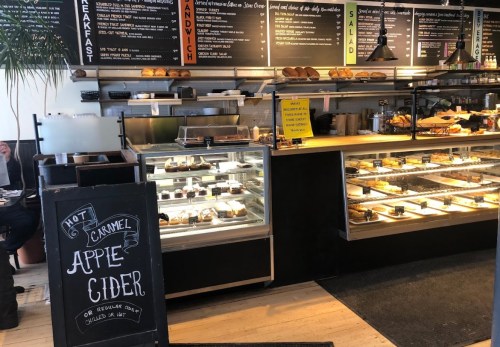 A cozy café display with pastries, a chalkboard sign for caramel apple cider, and a menu board overhead.