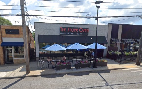 A bakery café with outdoor seating, blue umbrellas, and flower planters, located on a sunny street.
