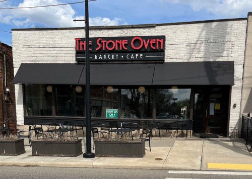 Exterior of The Stone Oven Bakery Cafe, featuring a sign, outdoor seating, and a sunny sky.