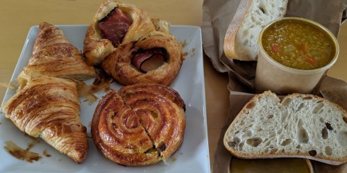 A plate of assorted pastries, including croissants and a cinnamon roll, alongside a bowl of soup and slices of bread.