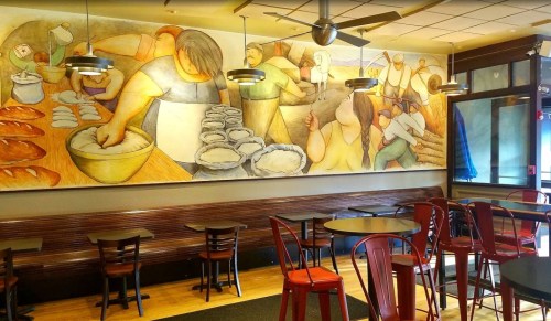 A cozy café interior featuring a mural of bakers and diners, with red chairs and wooden tables.