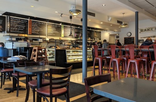A cozy café interior with wooden tables, red chairs, a display case, and a menu board in the background.
