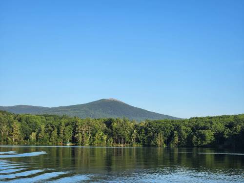 A serene lake reflects a clear blue sky and a mountain surrounded by lush green trees.