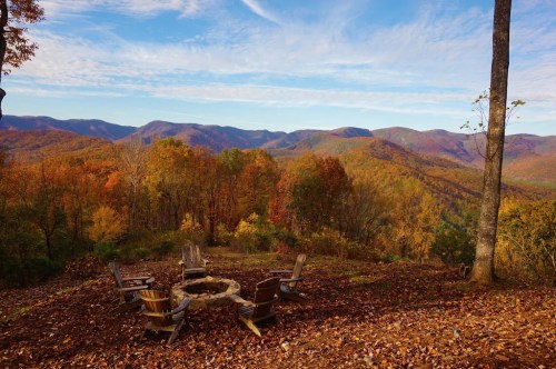 A scenic view of colorful autumn trees and mountains, with a circular stone fire pit and wooden chairs in the foreground.