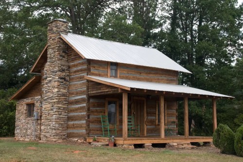 A rustic log cabin with a stone chimney and porch, surrounded by trees.