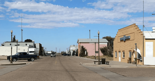 A quiet street in a small town, featuring buildings, parked vehicles, and a clear blue sky.