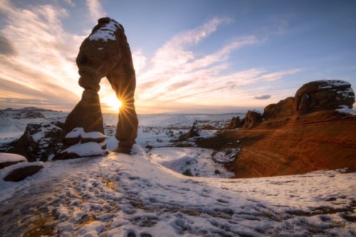 A stunning rock formation with a sunset backdrop, surrounded by snow-covered terrain and dramatic clouds.
