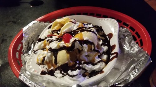 A dessert bowl filled with ice cream, whipped cream, chocolate sauce, and a cherry on top, served in a red basket.