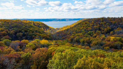 A scenic view of rolling hills covered in autumn foliage, overlooking a river under a partly cloudy sky.
