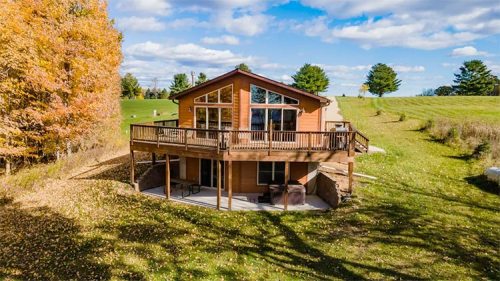 A spacious wooden house with large windows, surrounded by autumn foliage and a green lawn under a blue sky.