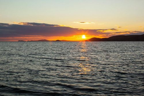 A serene sunset over calm waters, with silhouettes of distant hills against a colorful sky.