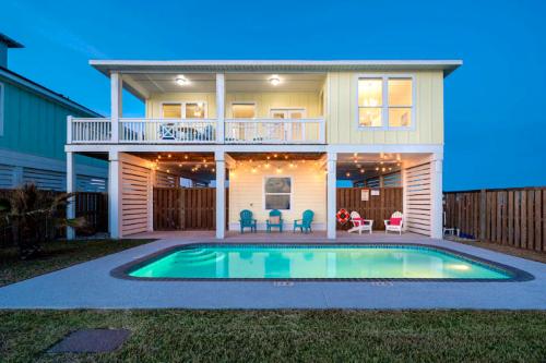 A two-story beach house with a pool, surrounded by string lights and chairs, set against a twilight sky.