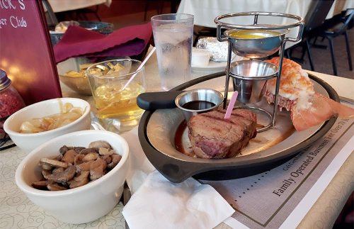 A table setting with a steak, lobster tail, mushrooms, and drinks, featuring a fondue pot in the center.