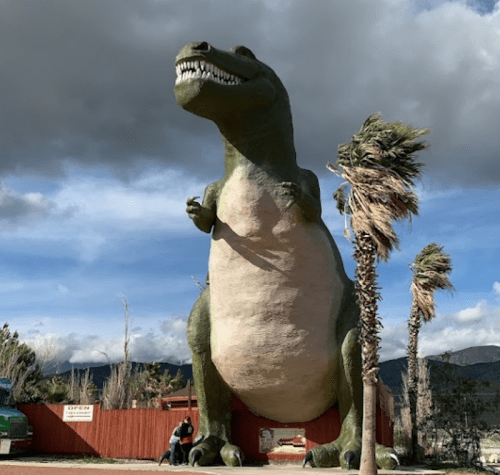 A large dinosaur statue stands in a park, with palm trees and cloudy skies in the background.