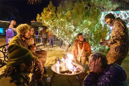 A group of people gathered around a campfire at night, surrounded by trees adorned with lights.