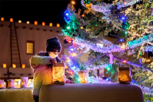 A child in a mask lights a paper lantern near a decorated Christmas tree at night. Colorful lights glow around.