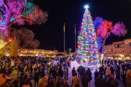 A festive crowd gathers around a brightly lit Christmas tree in a vibrant, colorful night scene.